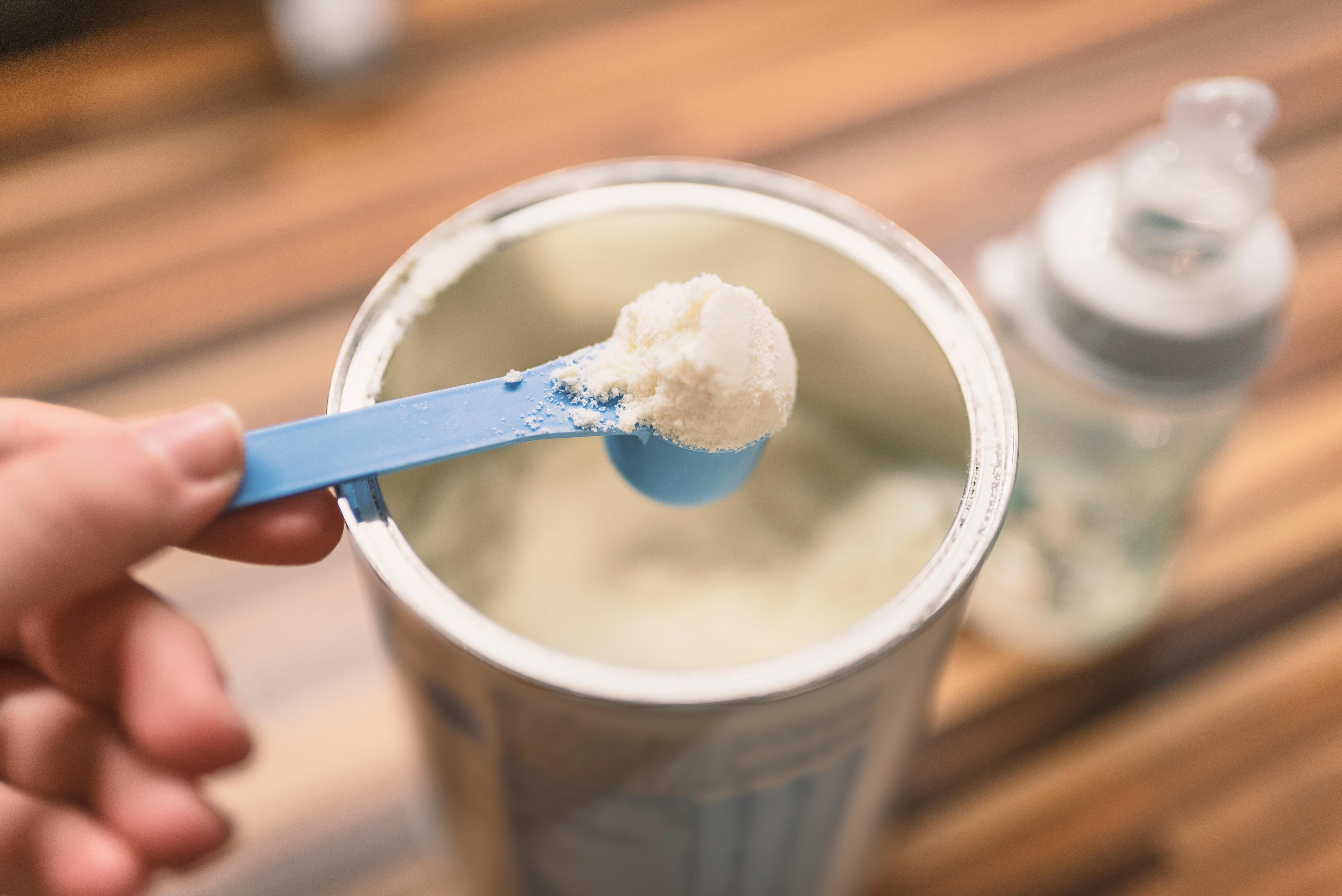 An adult's hand scooping infant formula from a can with a blurred baby bottle on the table.