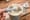 An adult's hand scooping infant formula from a can with a blurred baby bottle on the table.